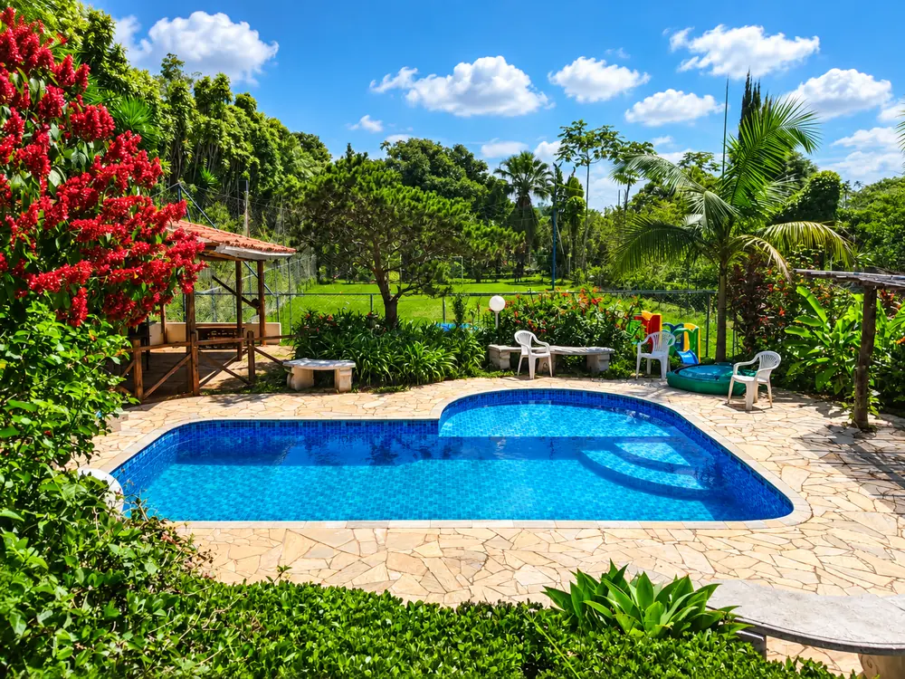 Piscina azul com deck de pedra, cercada por jardim florido e vista para o campo